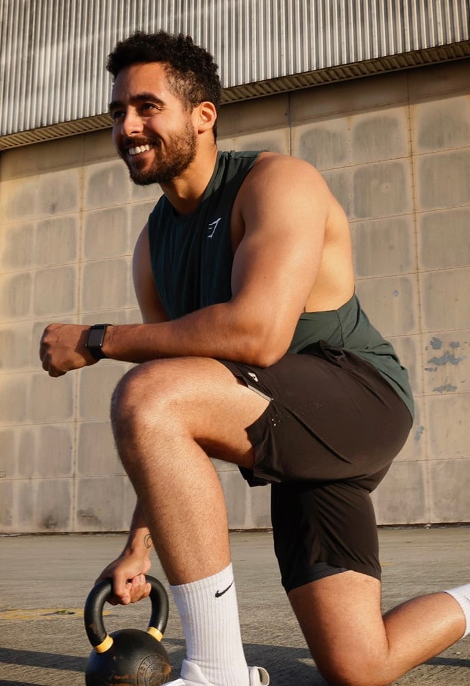 Man in athletic wear smiling while performing a lunge exercise with a kettlebell outdoors against an industrial wall